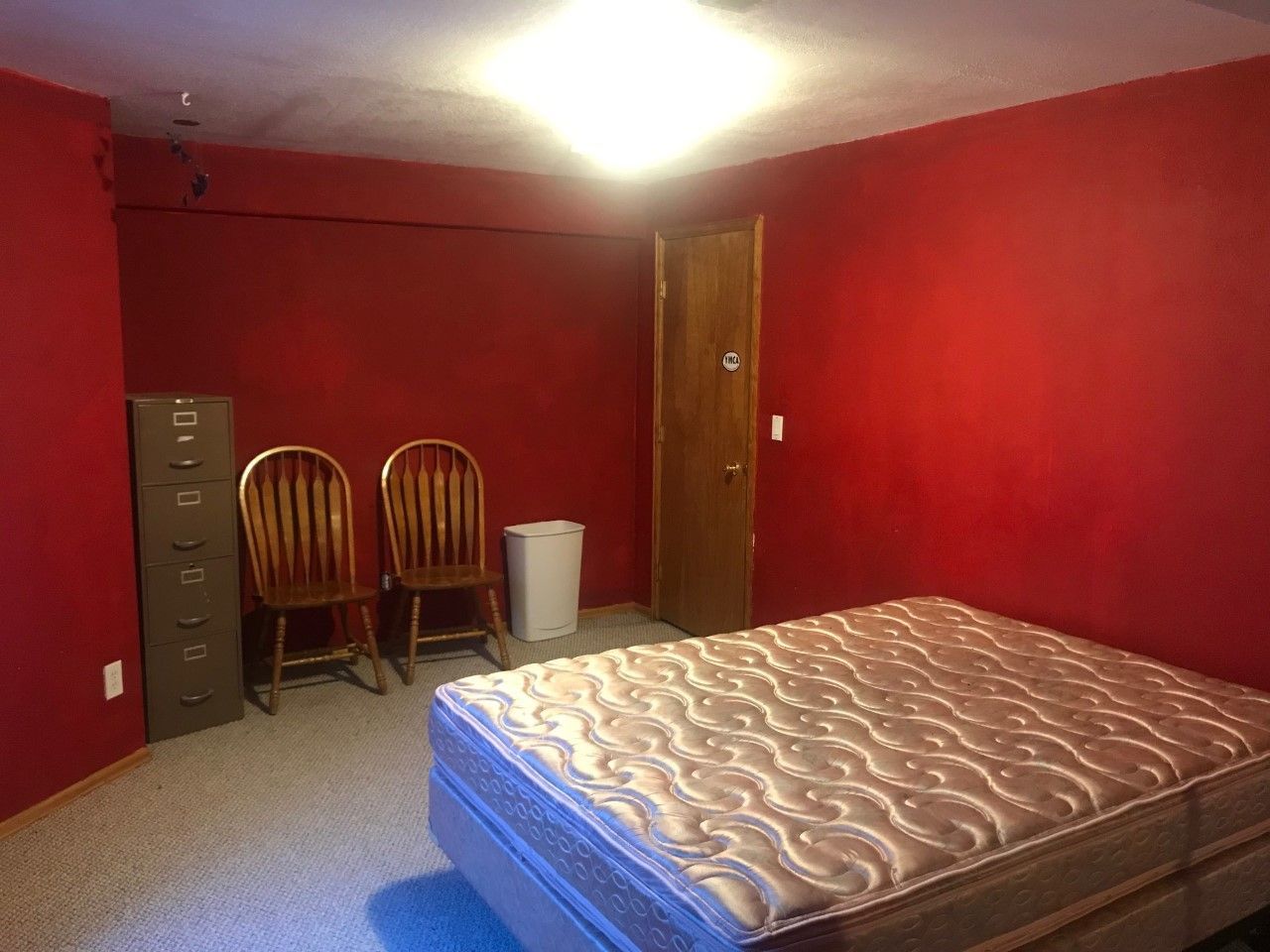 Red-walled room with mattress, two chairs, file cabinet, small fridge, and a closed door.