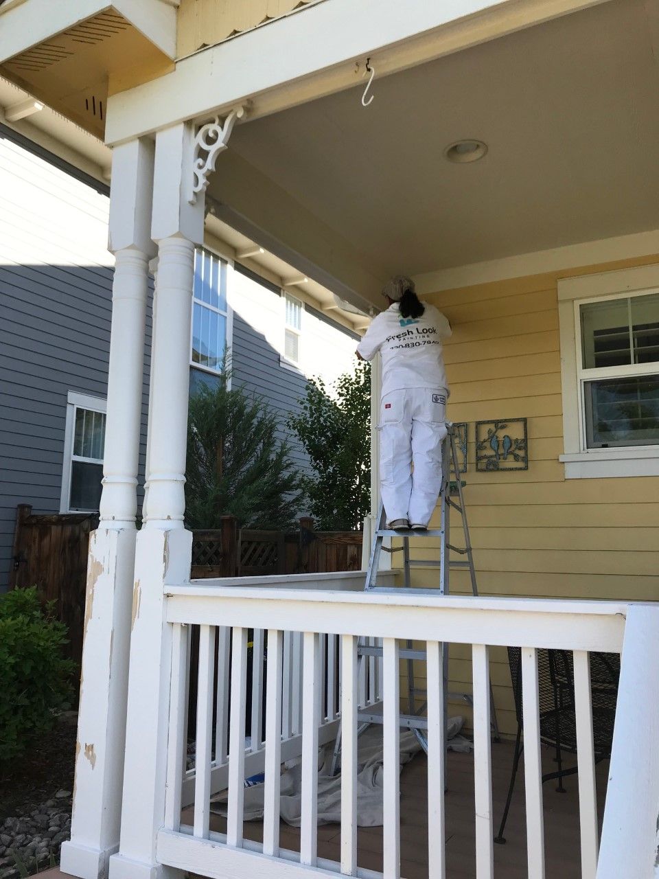 Person painting a porch ceiling, standing on a ladder. The porch is yellow with white trim.