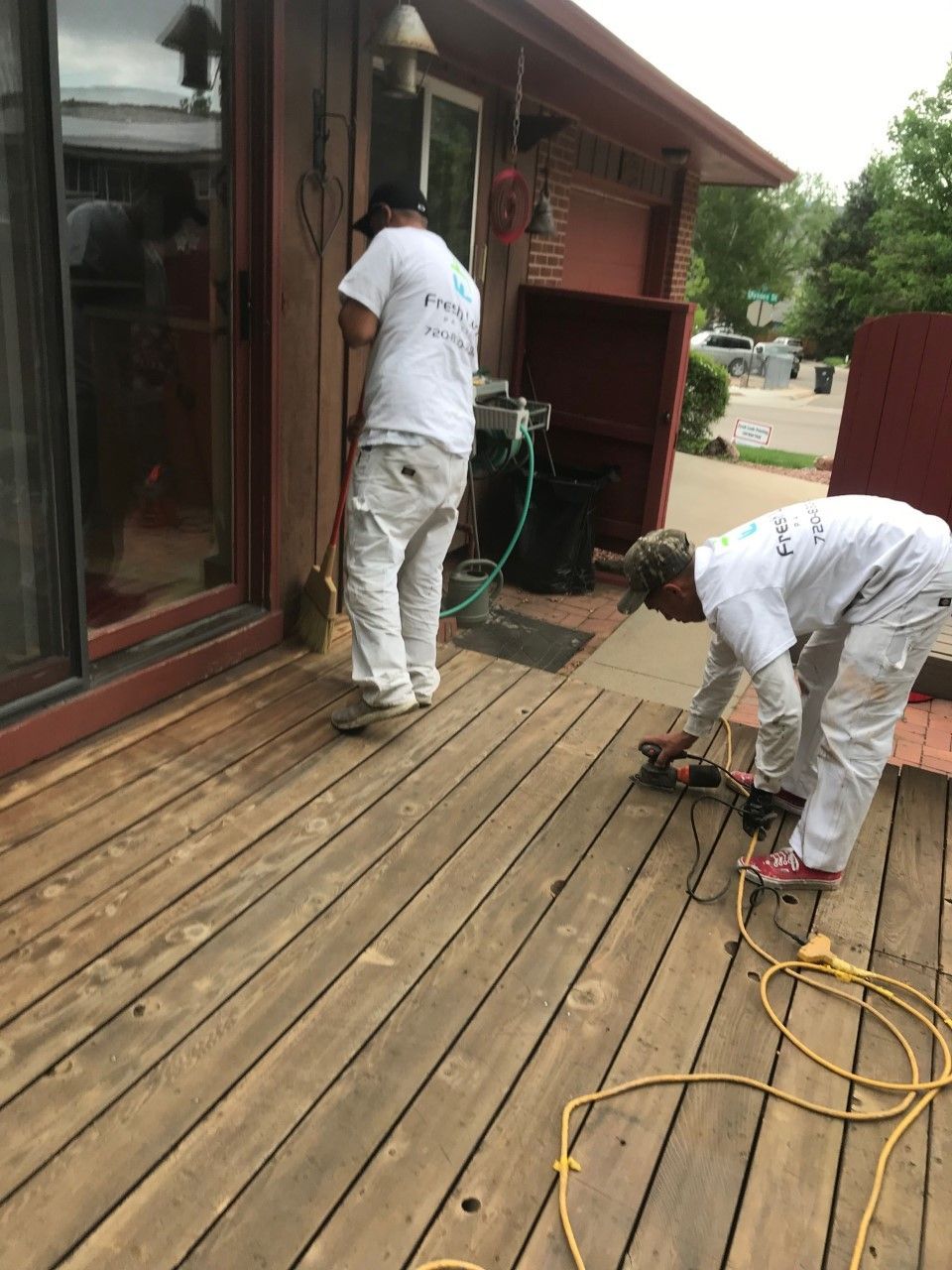 Two people sanding a wooden deck, preparing for staining. One sweeps, the other uses a sander.