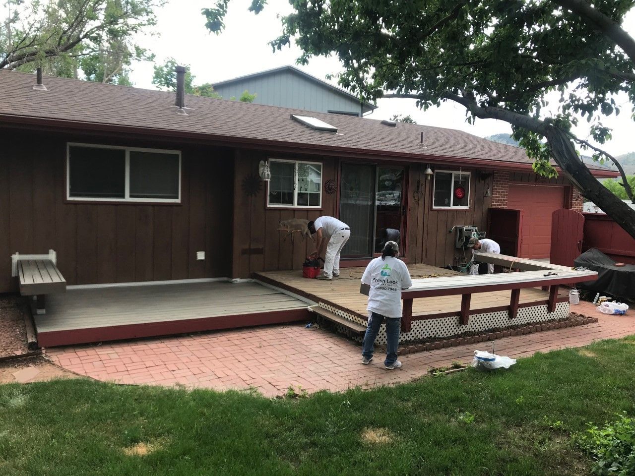 Workers paint a brown house exterior with a deck and brick patio.