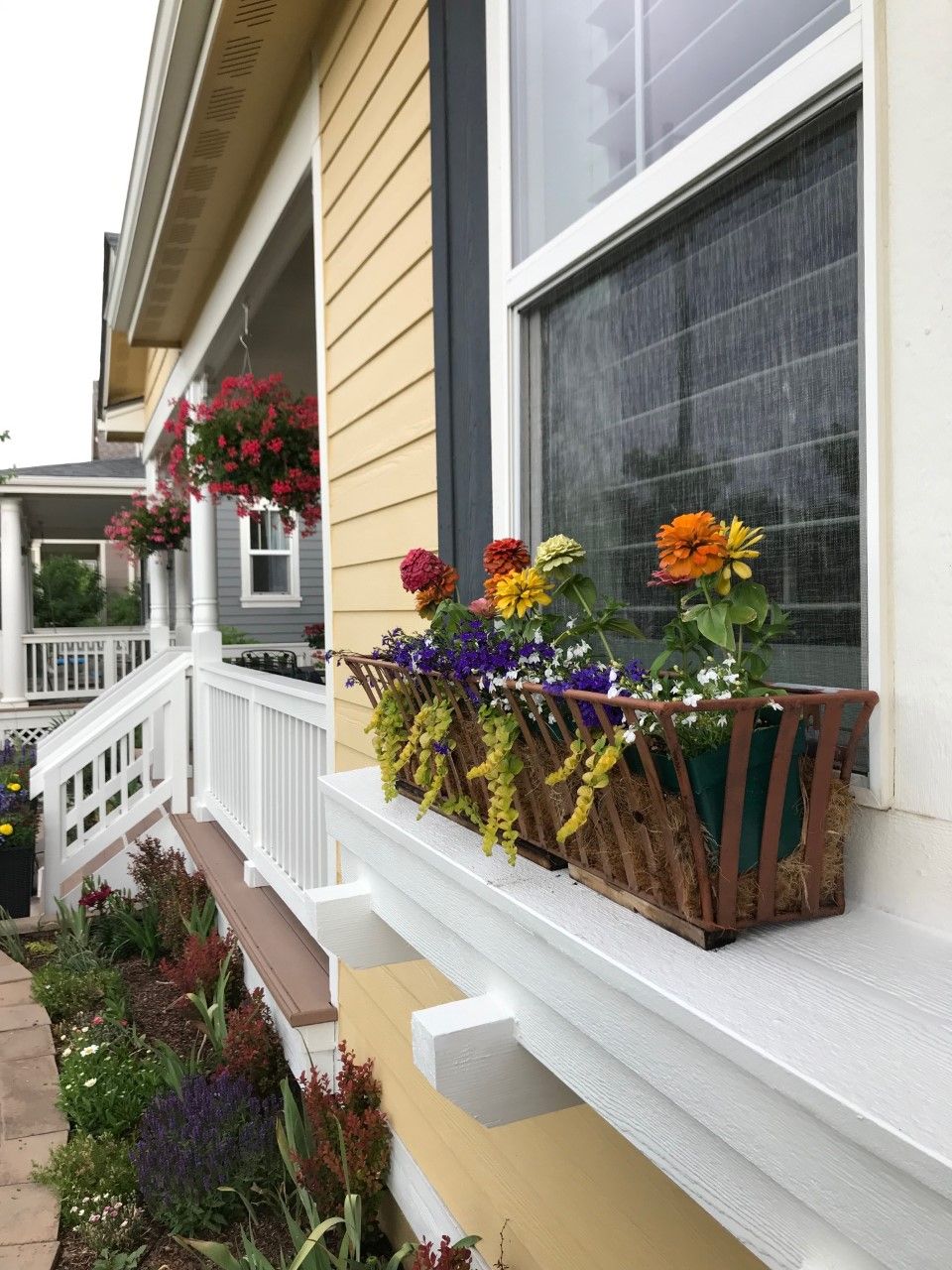 Yellow house with window box overflowing with colorful flowers, porch with hanging baskets.