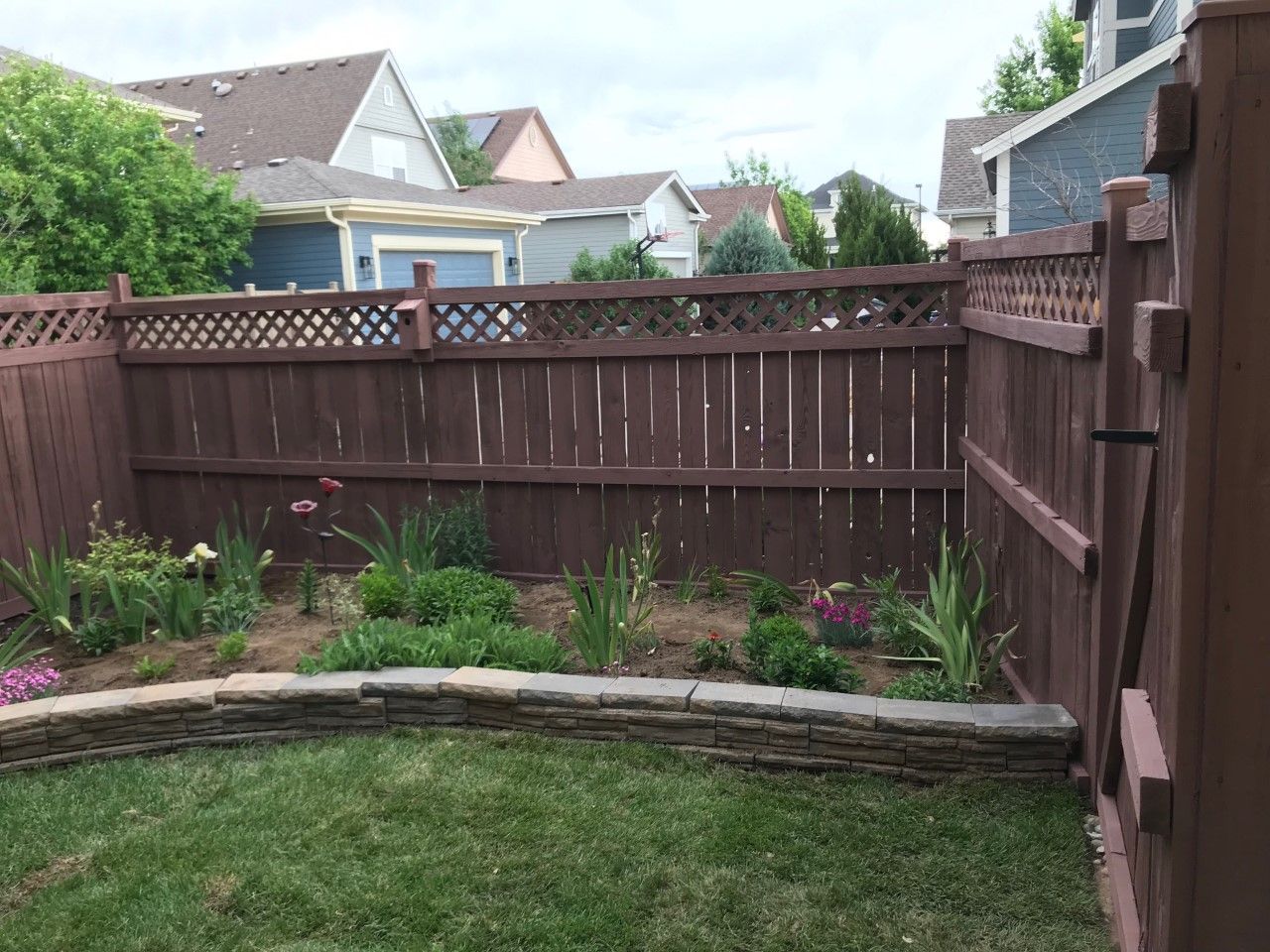 Brown wooden fence encloses a backyard with a flowerbed and green lawn.