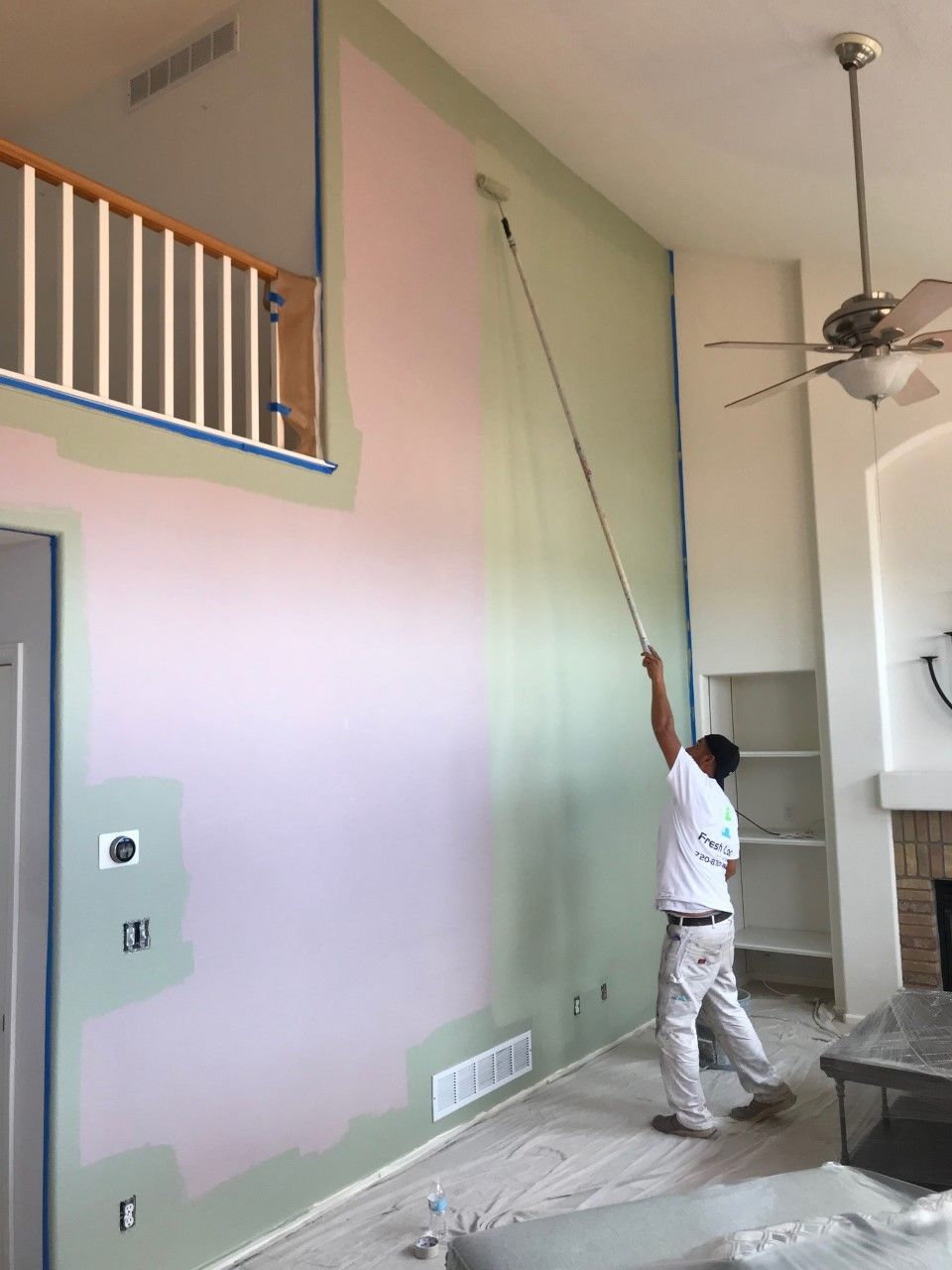 Painter using roller on high wall, two-tone paint. Blue tape protects edges. Interior setting, high ceiling.