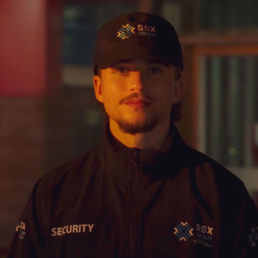 A man wearing a hat and a security jacket is looking at the camera.