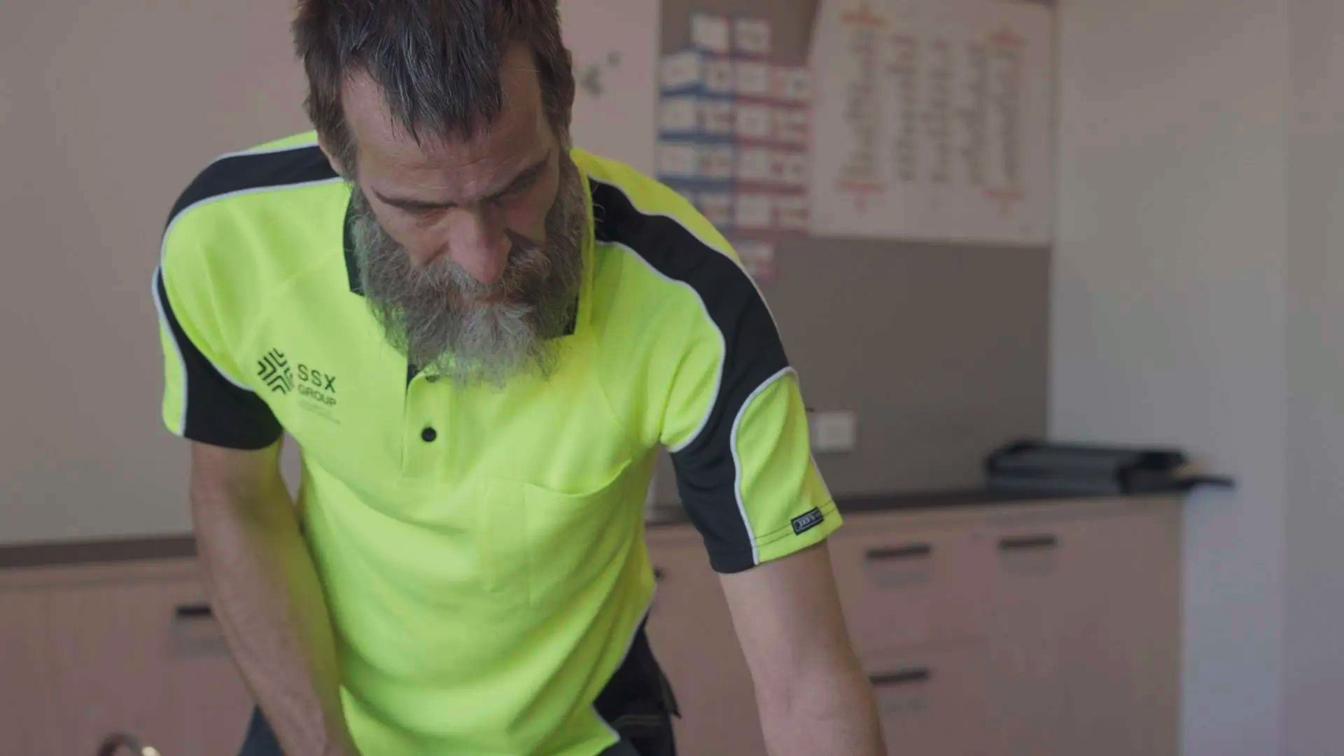 A man with a beard wearing a neon yellow shirt is standing in a kitchen.