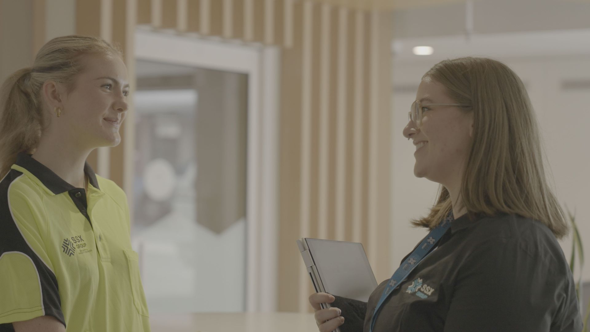 Two women are standing next to each other in a hallway talking to each other.