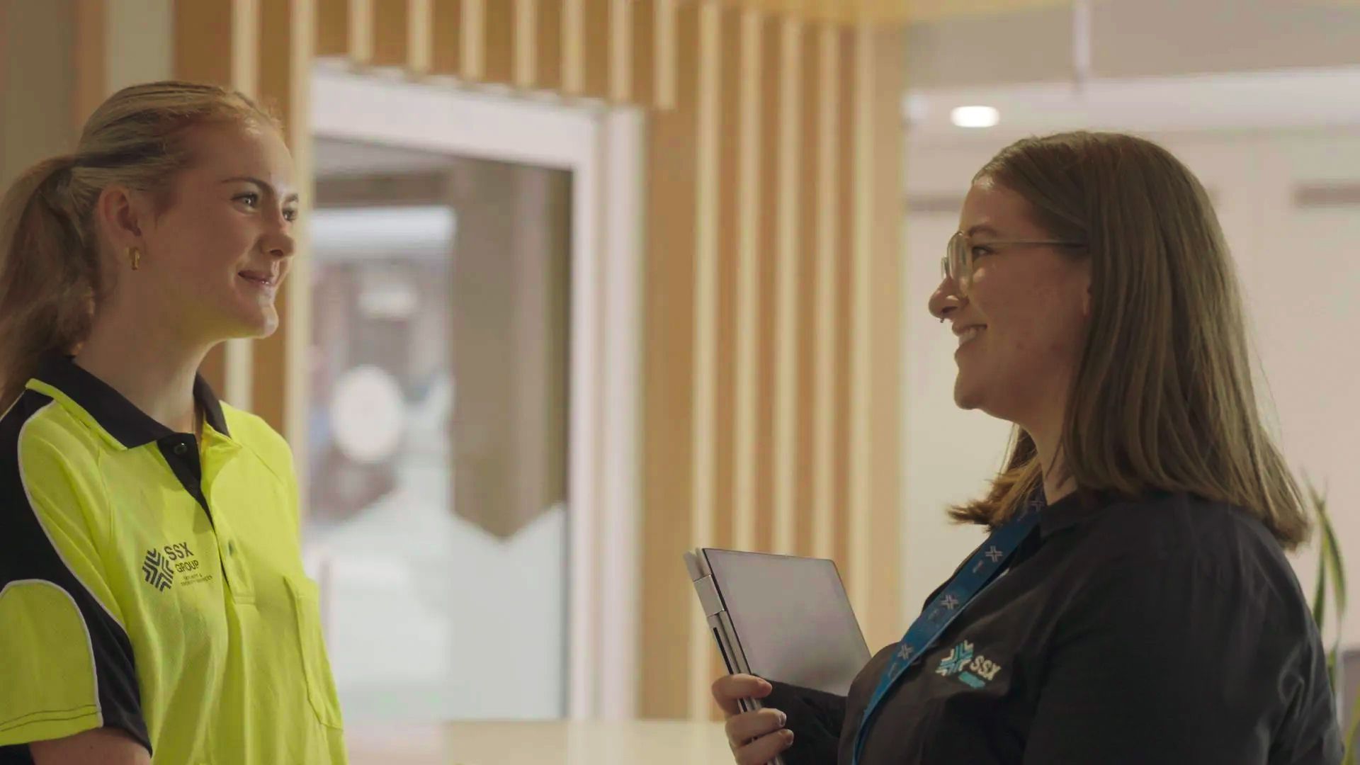 Two women are standing next to each other in a hallway talking to each other.