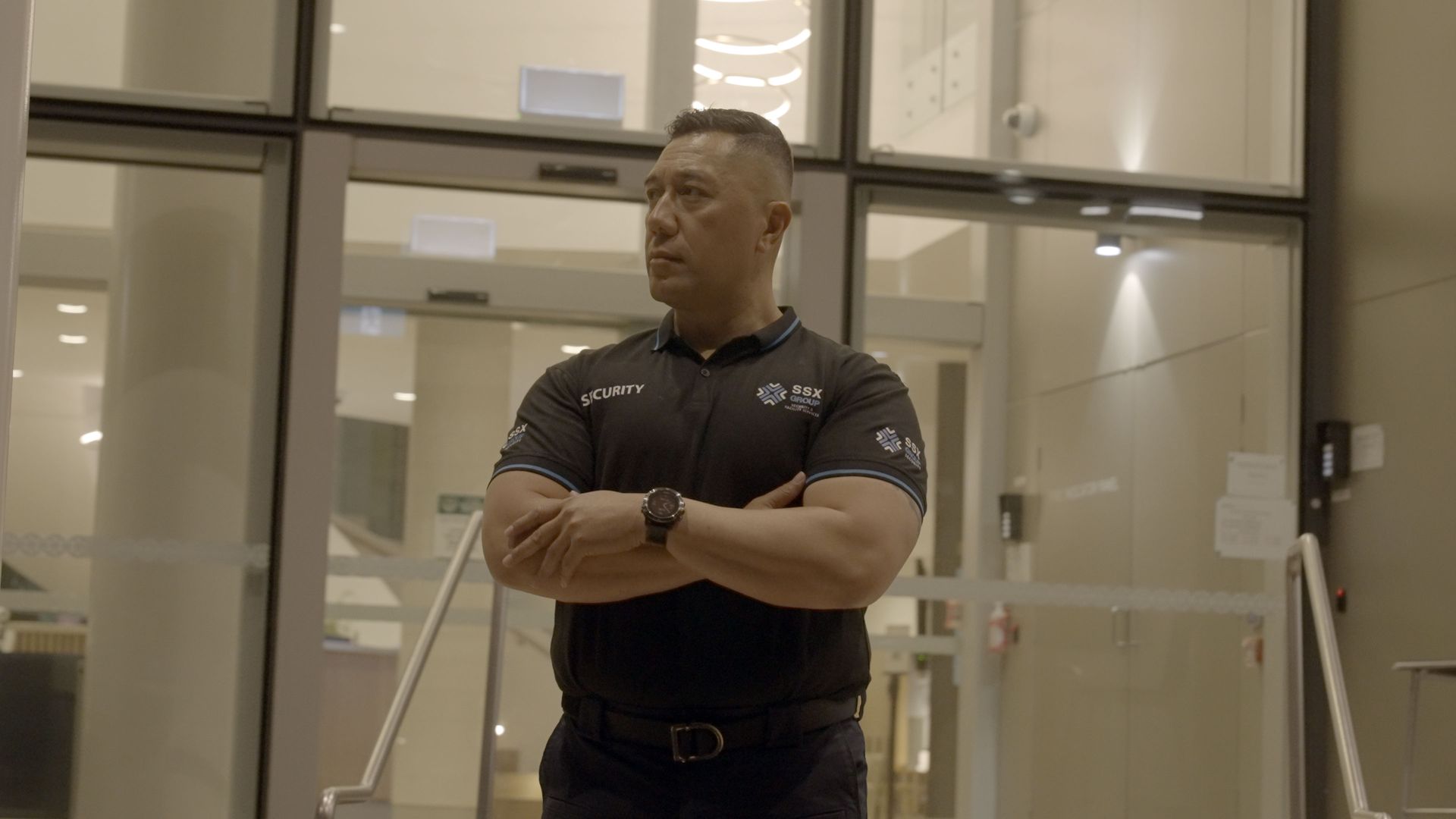 A security guard is standing in front of a glass door with his arms crossed.