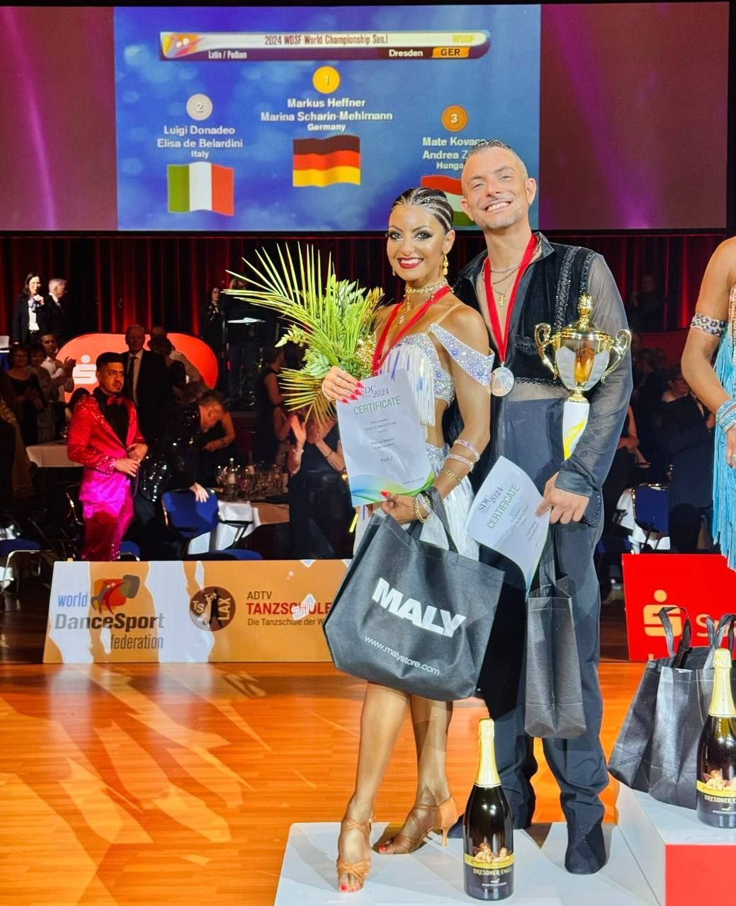 Una coppia di ballerini, uno dei quali tiene in mano un trofeo e dei fiori, festeggia su un podio con la bandiera italiana sullo sfondo.