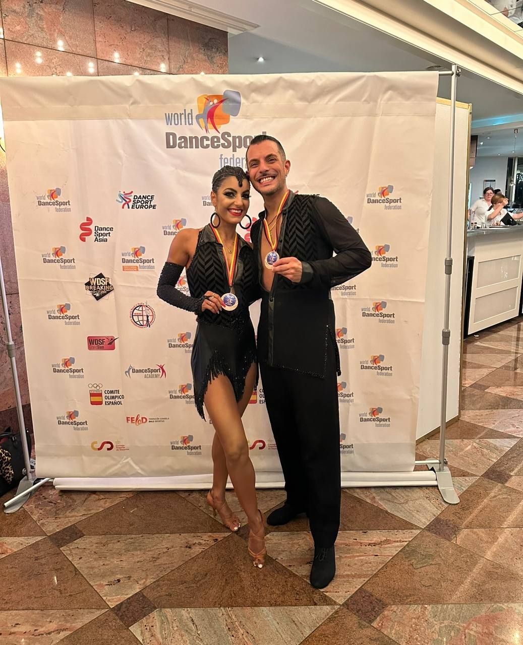 Una coppia di ballerini sorridenti, in abiti neri e medaglie, è in piedi davanti a uno striscione con i loghi. Sono in posa in un ambiente interno.