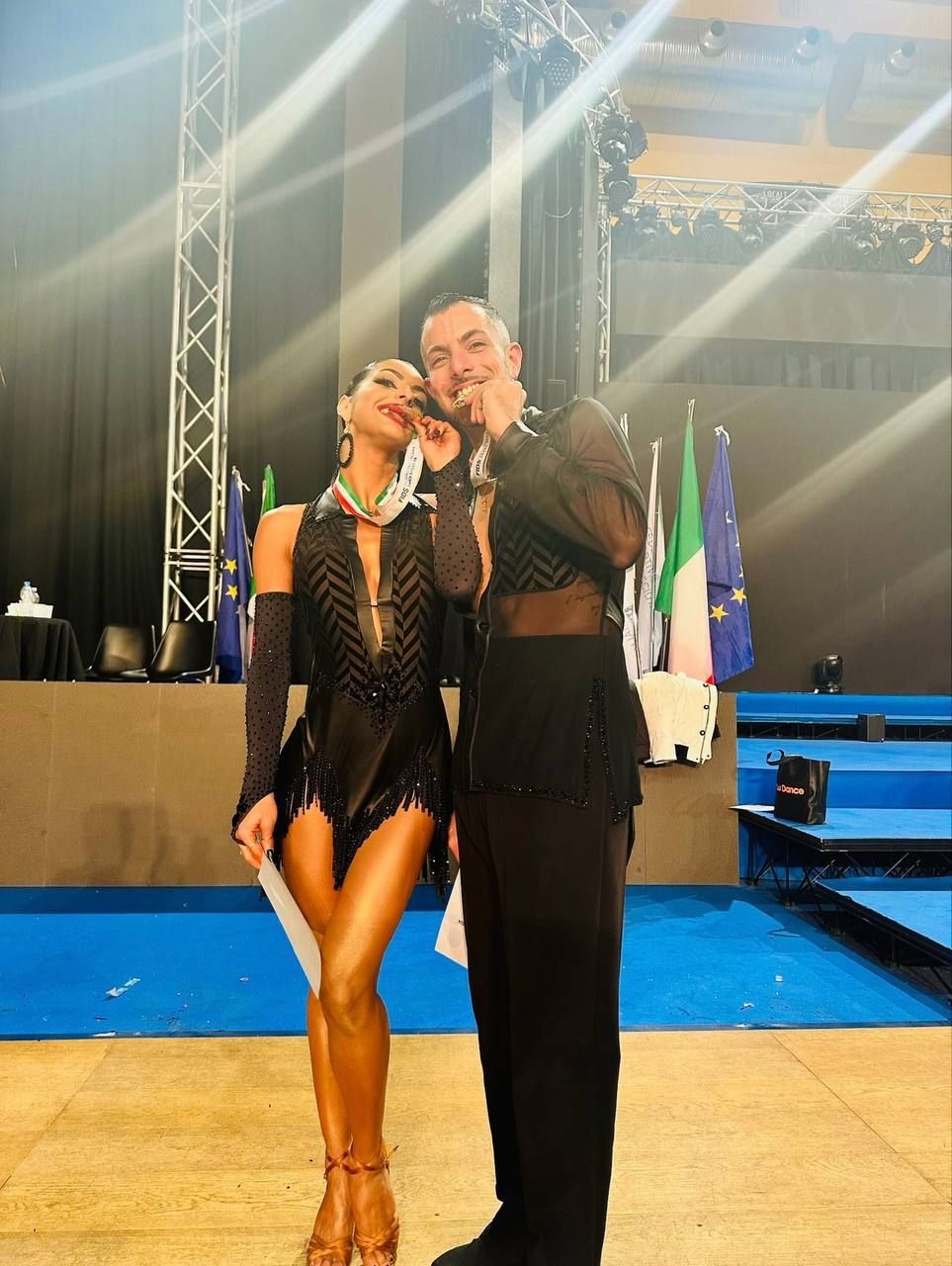 Una coppia di ballerini, un uomo e una donna, in abito nero formale, posano su una pista da ballo. La donna si tocca il viso mentre l'uomo si indica la bocca, entrambi sorridenti.