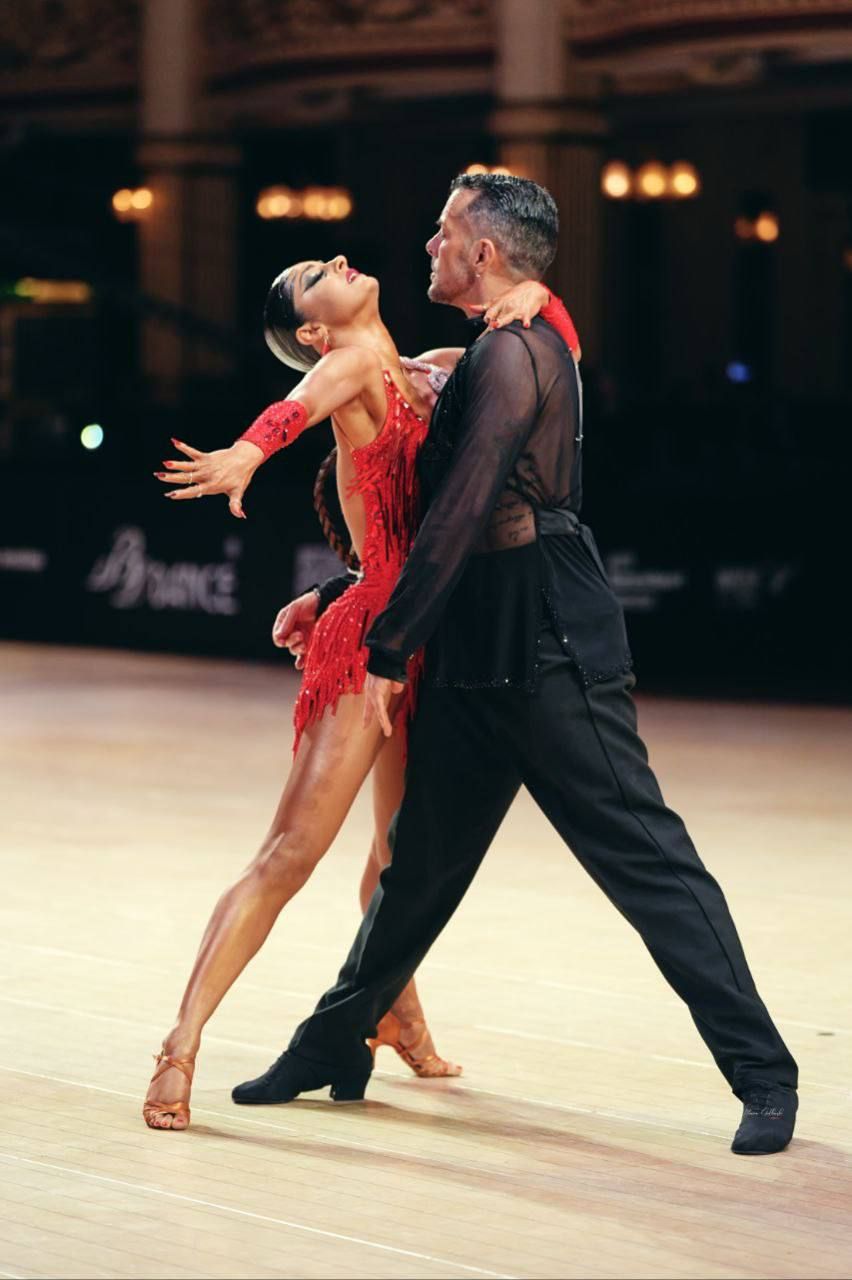 Coppia che esegue una danza passionale. La donna in abito rosso è appoggiata allo schienale, con le braccia tese, mentre l'uomo in abito nero la guarda.