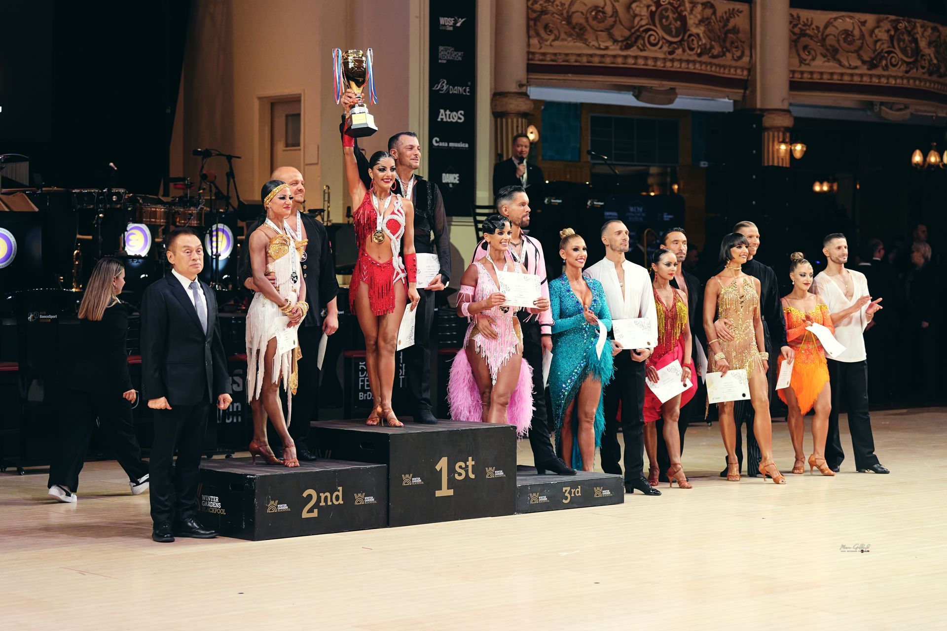 I vincitori di una gara di danza su un podio. Una coppia tiene in alto un trofeo, circondata da altri ballerini, giudici e da uno sfondo scenico.