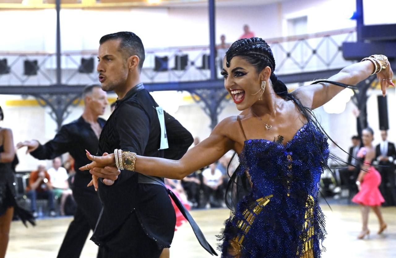 Una donna con un abito da ballo blu e un uomo vestito di nero si esibiscono in balli da sala con espressioni intense durante un evento.