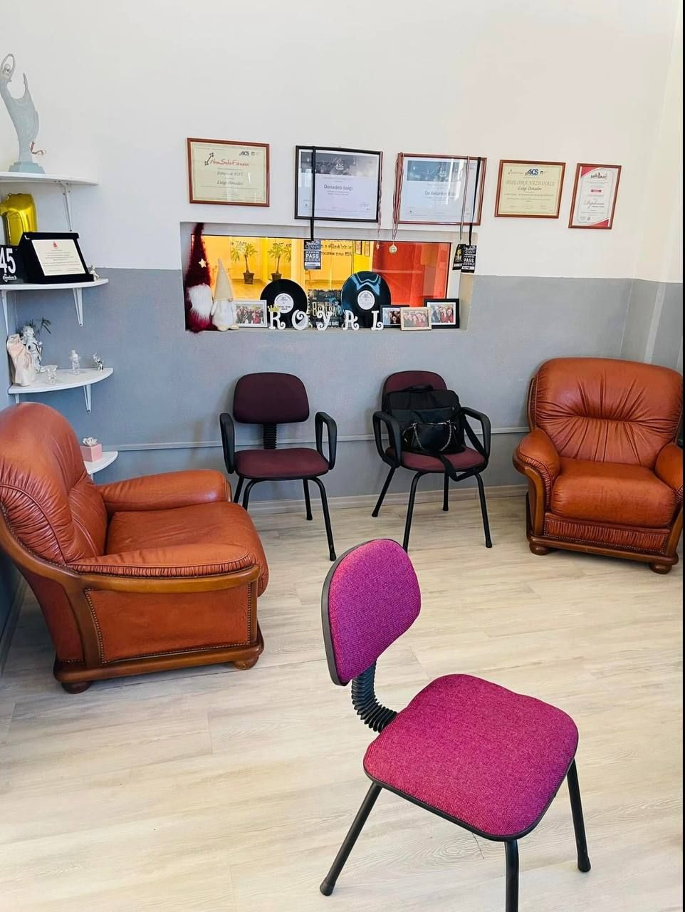 Sala d'attesa con mobili spaiati: due poltrone in pelle marrone, tre sedie scure e una sedia rosa. Premi appesi al muro.