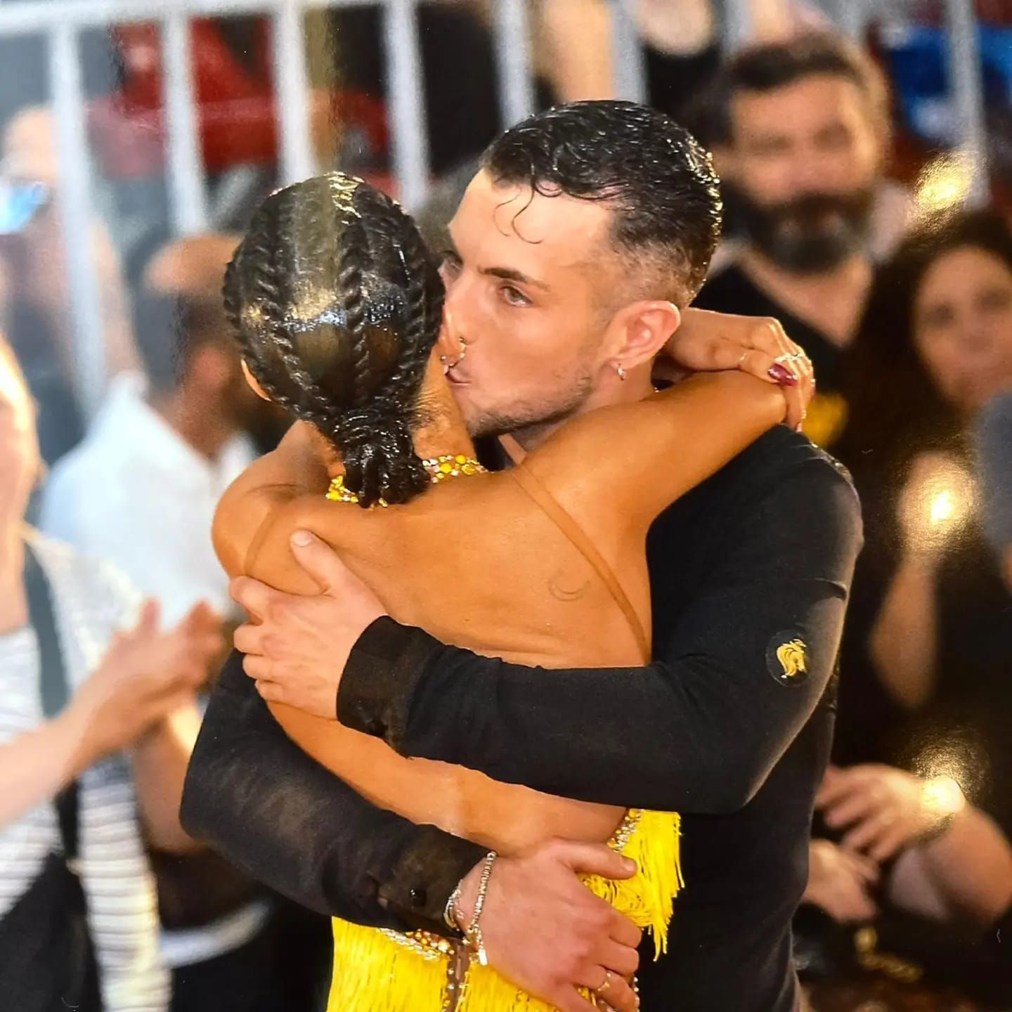 Un uomo bacia la guancia di una donna mentre la abbraccia. Sono su una pista da ballo, circondati da altre persone.