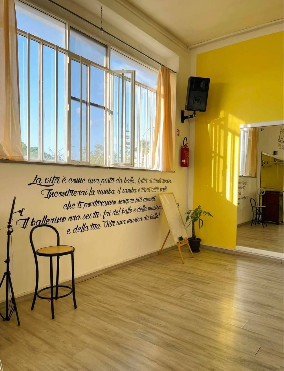 Interno di uno studio di danza con pareti gialle, ampie finestre e pavimento in legno. Nella stanza sono presenti uno sgabello, un cavalletto e una pianta.