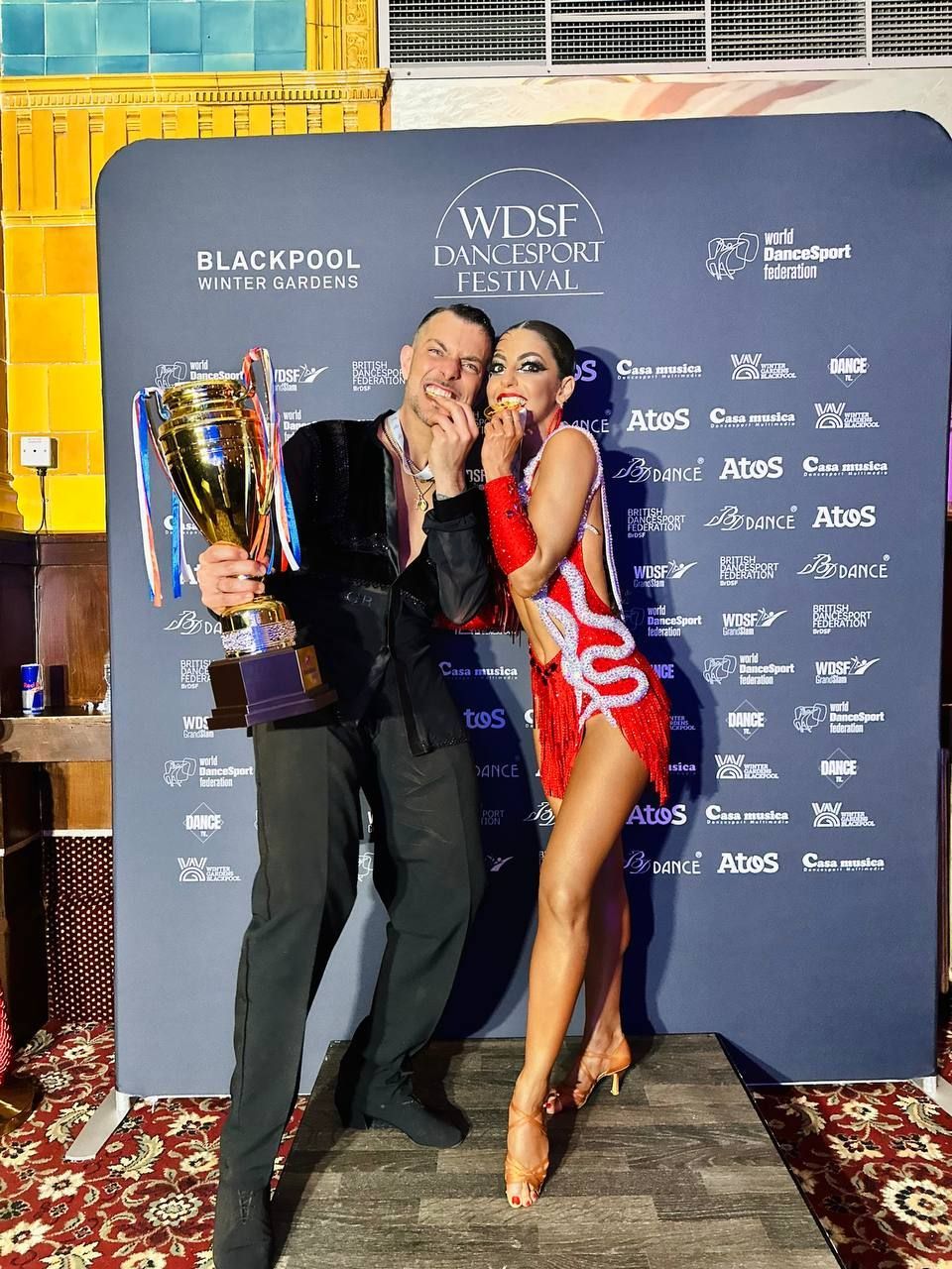 Una coppia di ballerini, un uomo e una donna, festeggiano tenendo in mano un trofeo davanti a uno sfondo: l'uomo indossa una giacca nera e la donna un vestito rosso.