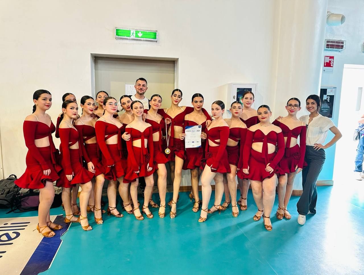 Un gruppo di ballerini in costumi rossi coordinati posa per una foto dopo una gara. Si trovano in un ambiente al chiuso, con una porta e alcuni spettatori visibili.