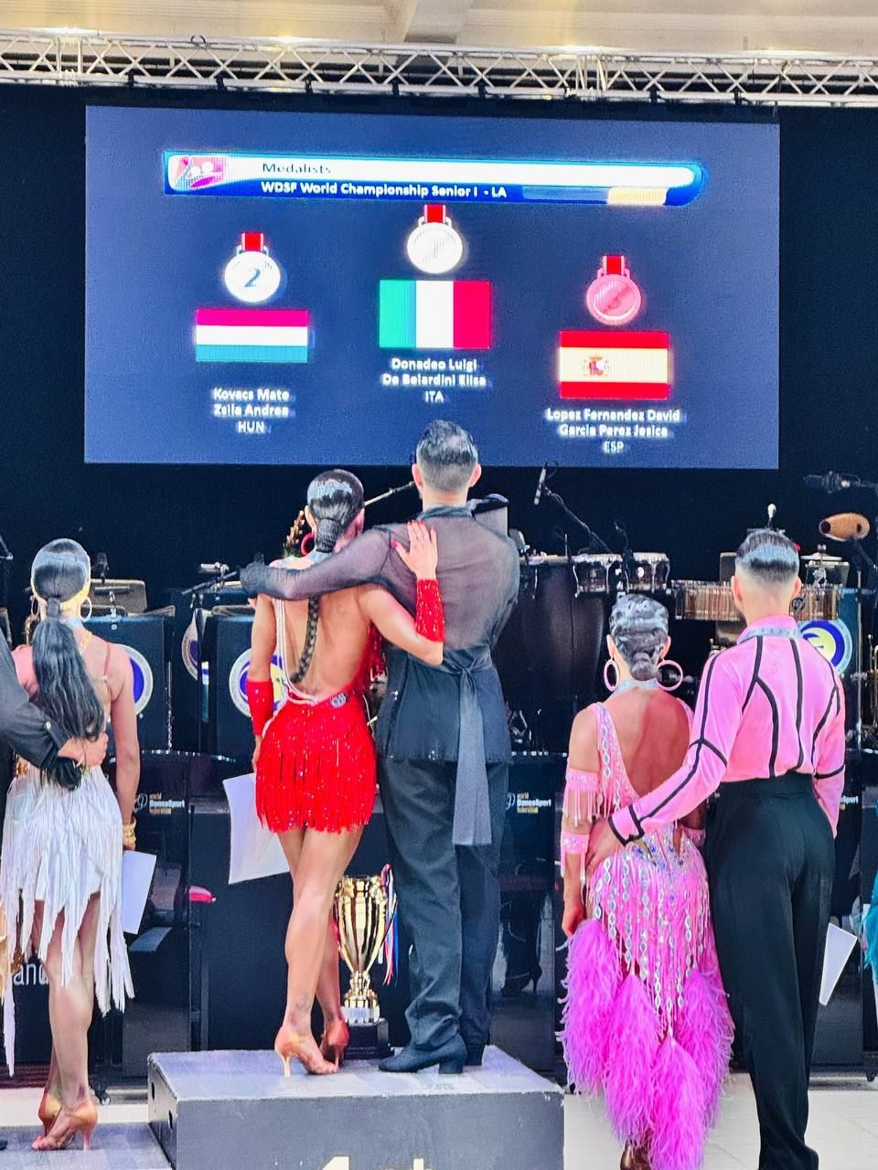Ballerini di ballo da sala su un podio, celebrano una gara. Le bandiere di Ungheria, Italia e Perù rappresentano i primi tre vincitori visualizzati sullo schermo.