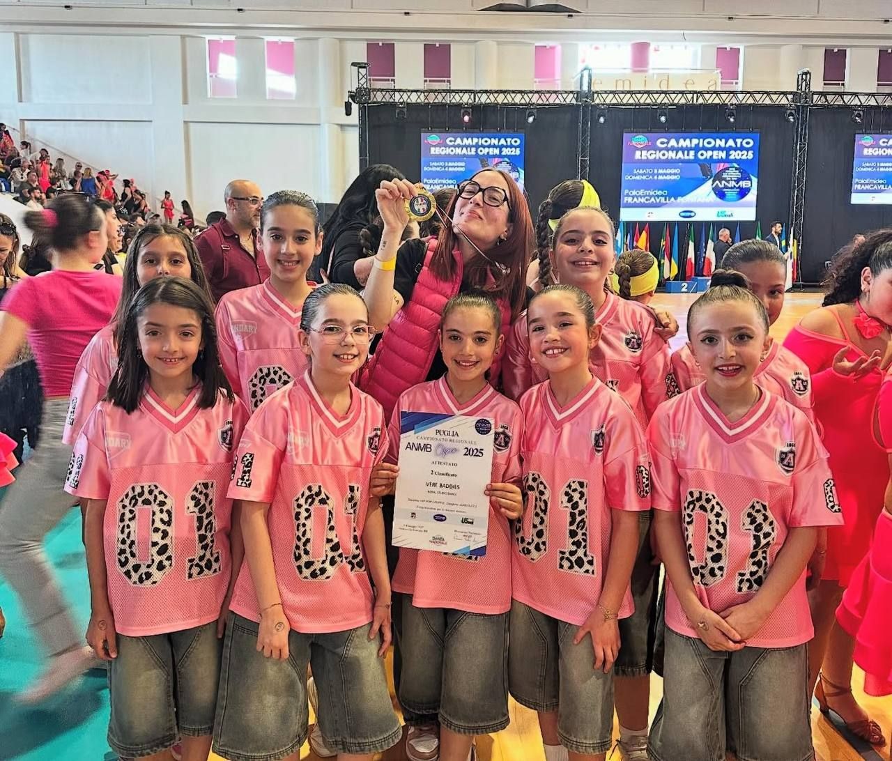 Una squadra di ballo composta da giovani ragazze in maglie rosa e pantaloncini di jeans festeggia con un attestato e una medaglia durante una gara di ballo.