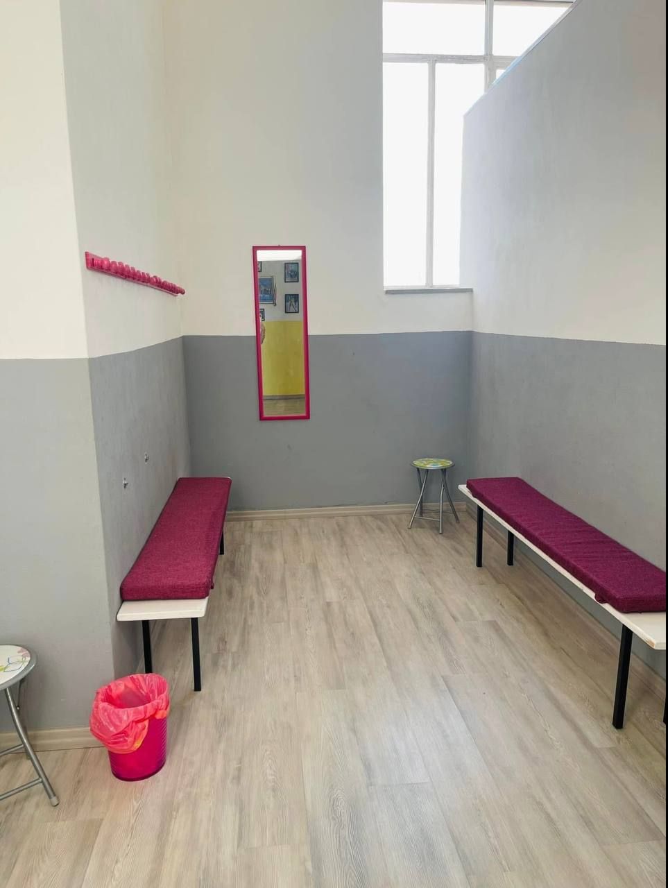 Uno spogliatoio con un attaccapanni rosa, panche, uno specchio e un cestino. Pareti grigie e bianche, pavimenti in legno chiaro.
