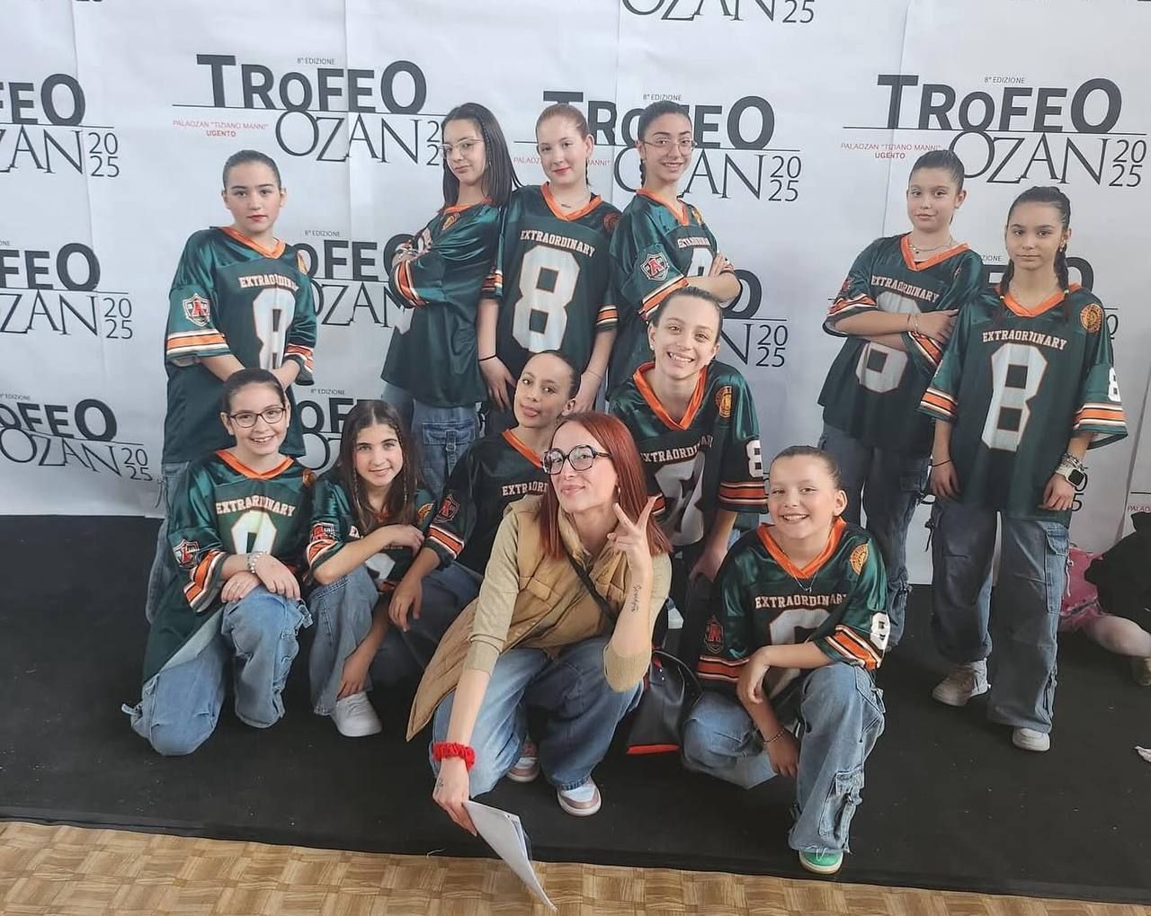 Un gruppo di giovani ballerini con maglie verdi e nere posa per una foto durante una gara di ballo con uno striscione con la scritta 