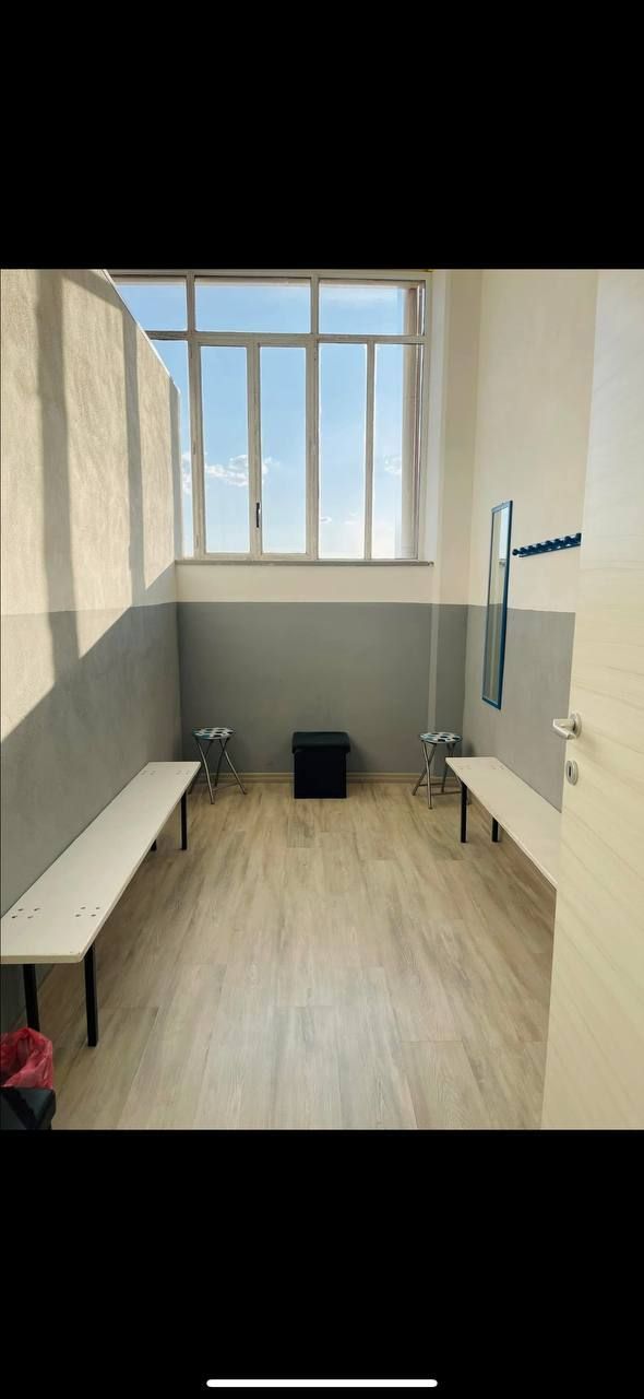 Uno spogliatoio con due panche, una finestra e un piccolo sgabello nero al centro. Le pareti sono grigie e bianche e il pavimento è in legno.