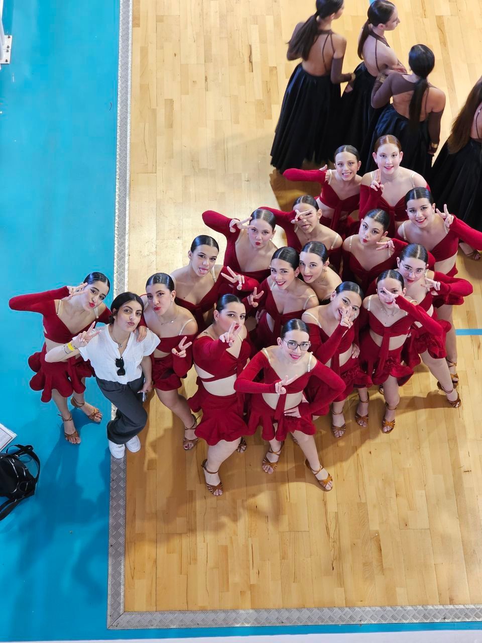 Un gruppo di giovani danzatrici in costume rosso posa su un pavimento di legno. Una donna con una camicia bianca è in piedi lì vicino.