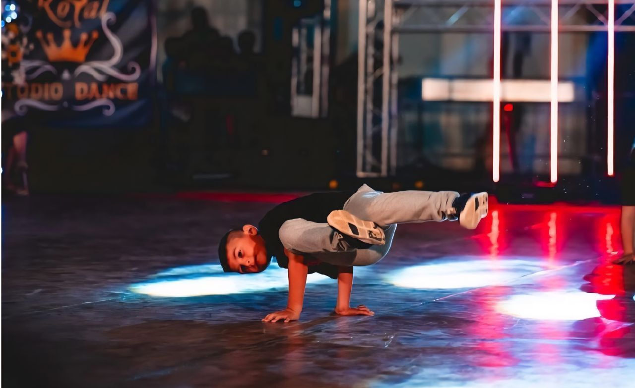 Un ragazzino balla la breakdance su un palco, in equilibrio su una mano e con le gambe sollevate. Luci rosse e blu illuminano la scena.