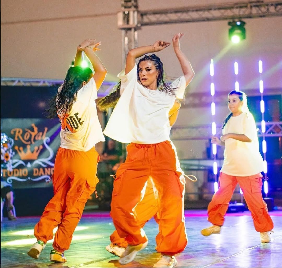 Tre ballerine in pantaloni arancioni e camicie bianche si esibiscono su un palco illuminato da luci colorate.