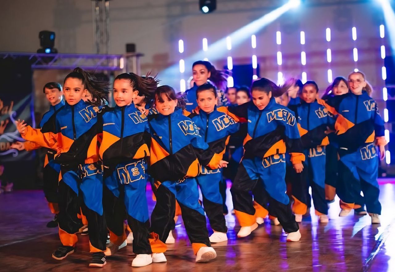 Un gruppo di danza si esibisce sul palco indossando tute blu e arancioni. I ballerini si muovono con espressioni concentrate sotto le luci del palco.