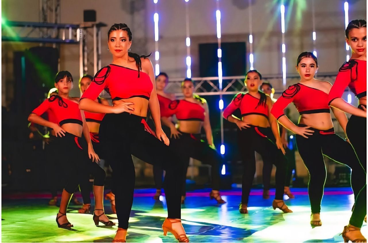 Un corpo di ballo femminile, vestito con abiti coordinati rossi e neri, esegue una danza sullo sfondo di un palco.