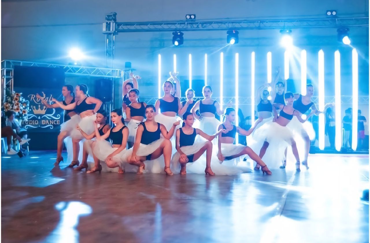 Ballerini con top neri e gonne bianche si esibiscono su un palco illuminato da luci blu.