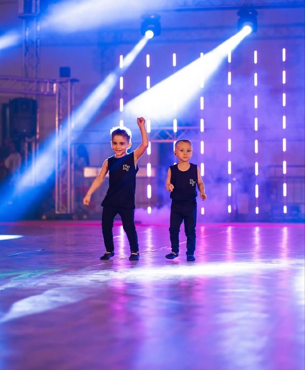 Due ragazzi in abiti neri ballano su un palco illuminato da luci blu e viola. Uno dei ragazzi ha un braccio alzato.
