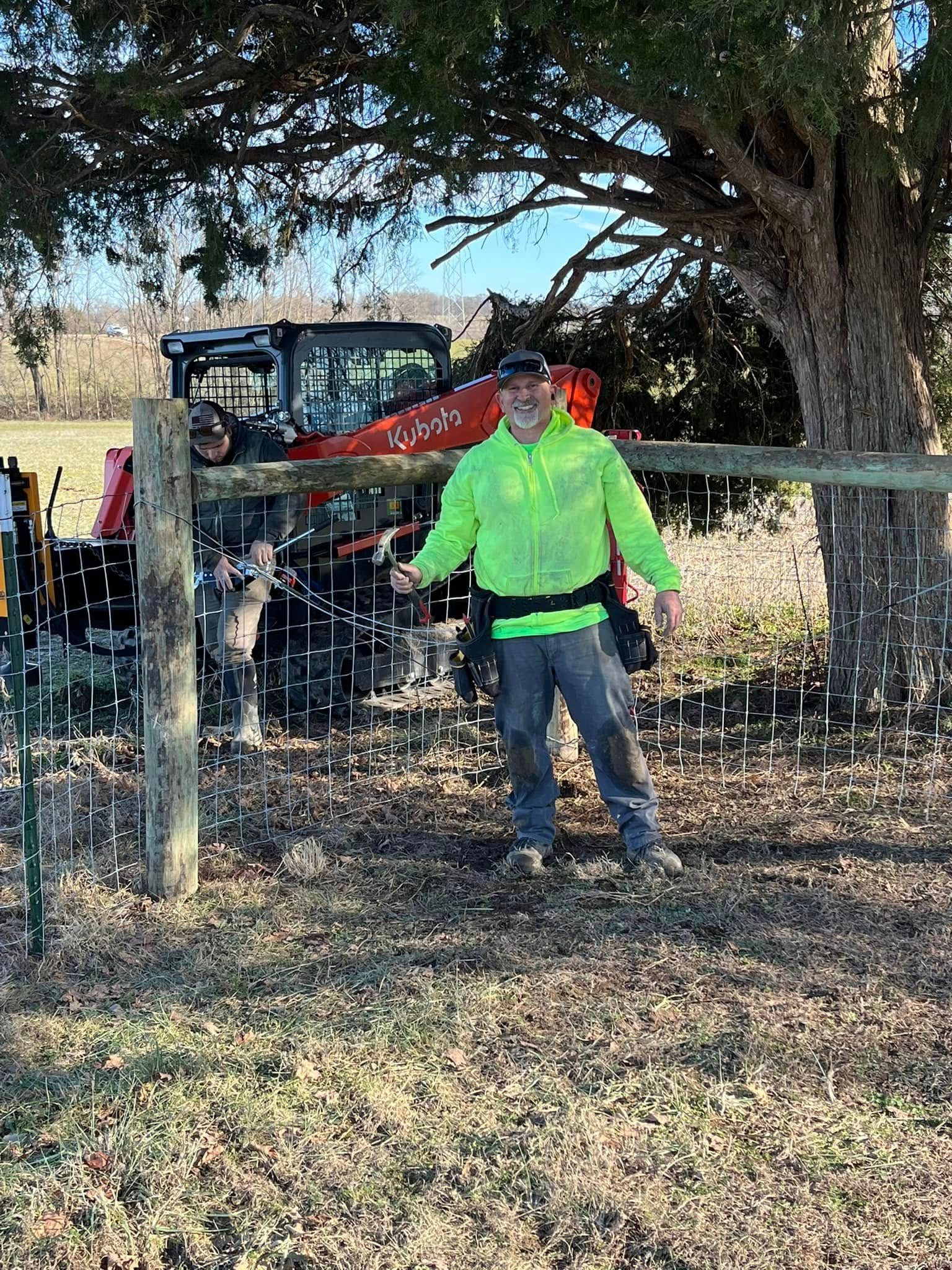 A man in a neon green jacket is standing next to a fence in a field.