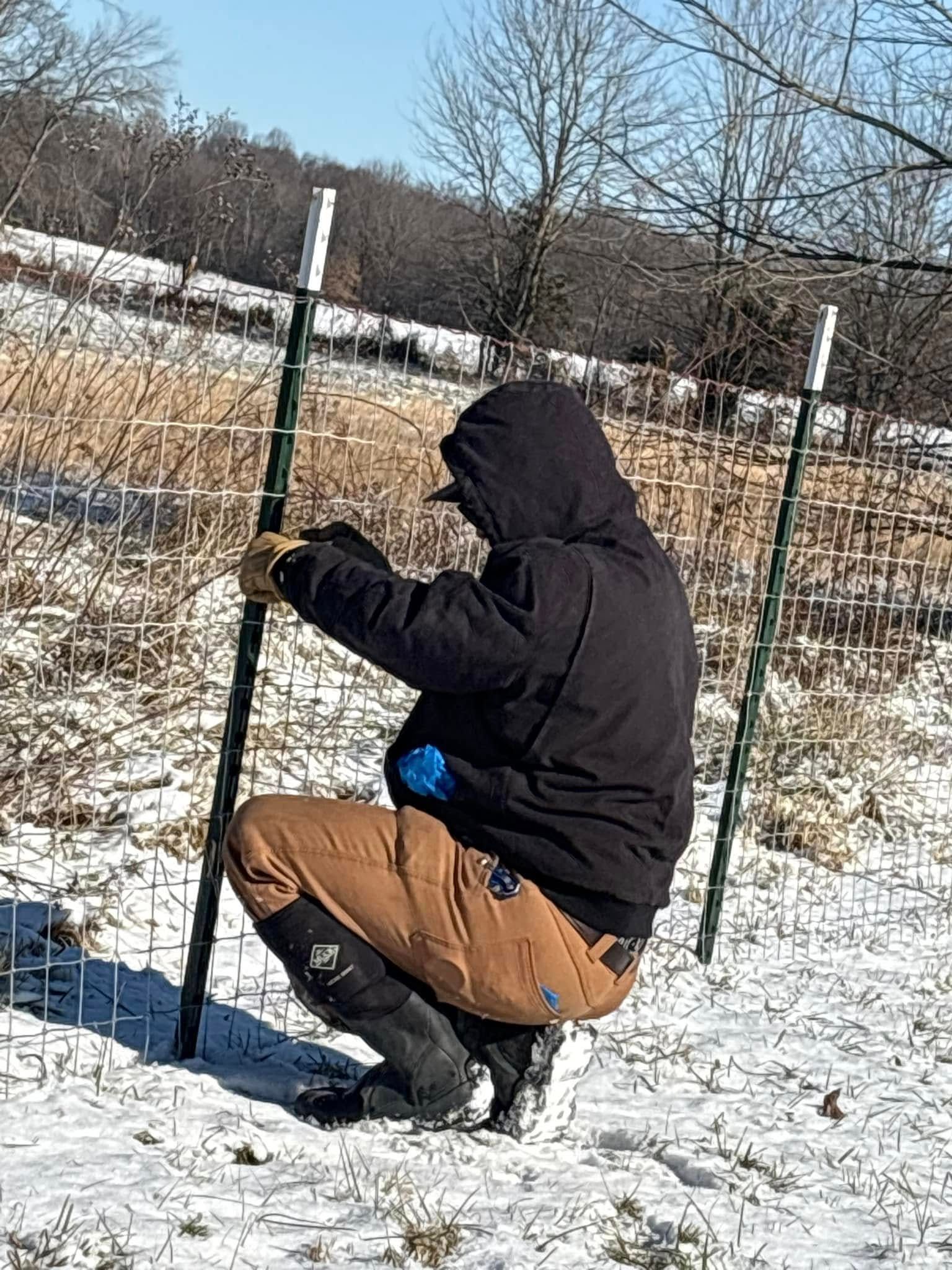 A man is kneeling down in the snow holding a fence post.