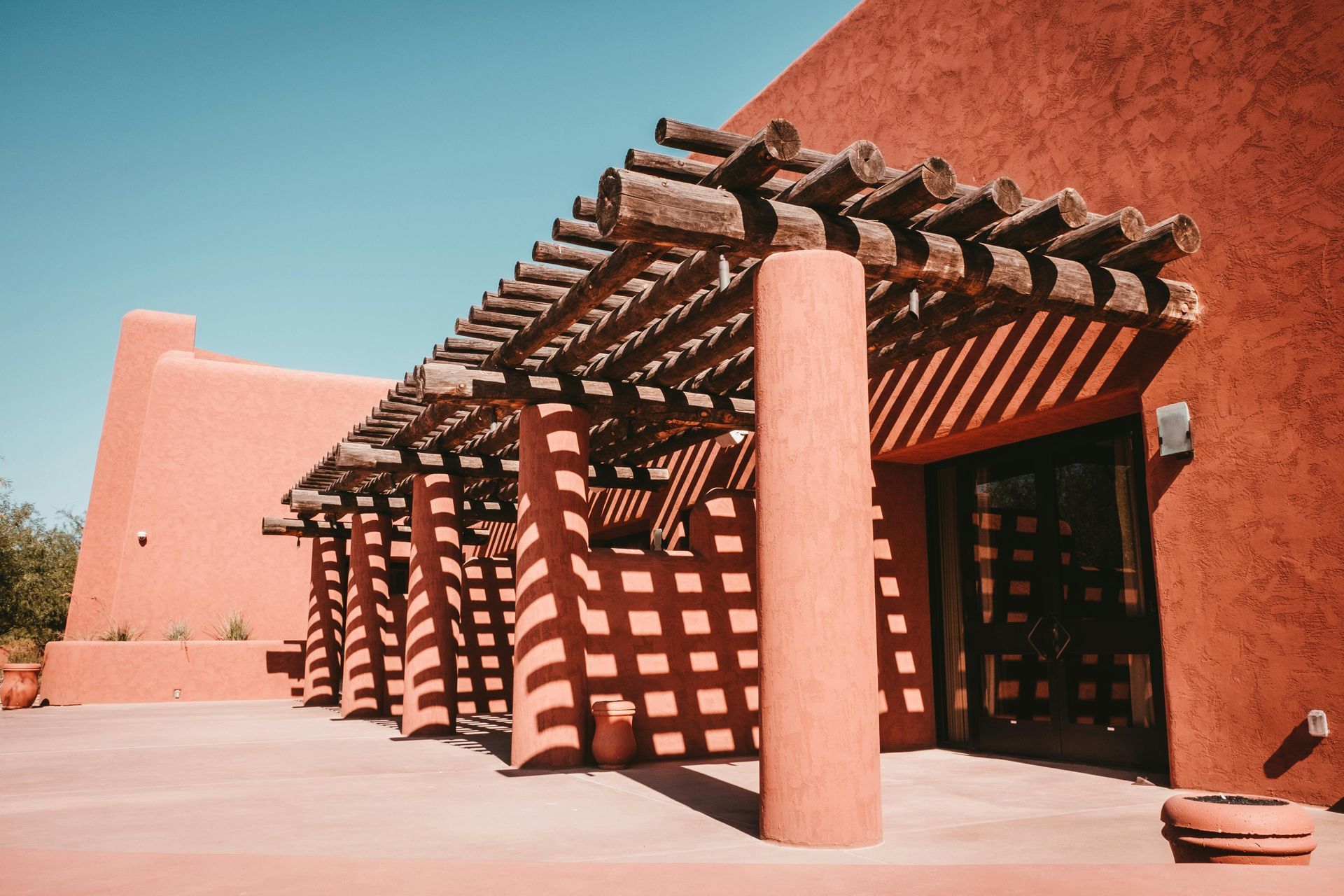 Red adobe building with wooden pergola, creating shadows in the sunlight.