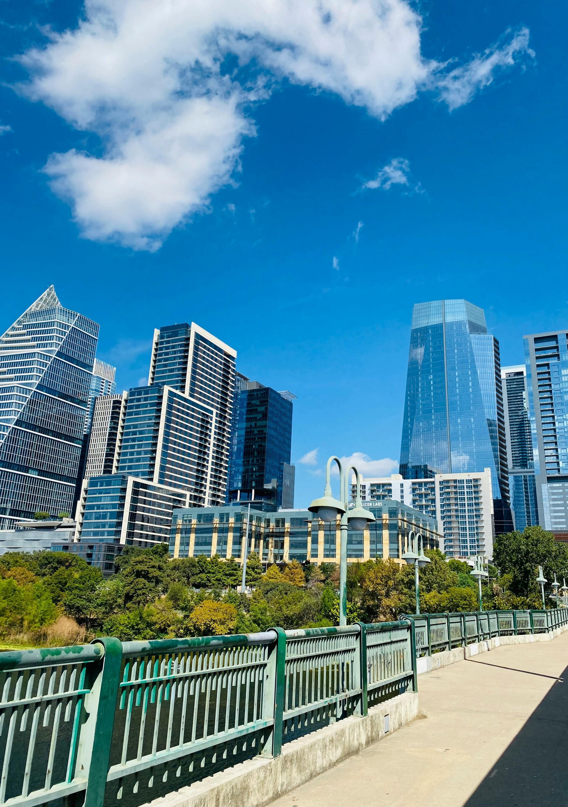 City skyline with modern buildings under a bright blue sky, viewed from a pedestrian bridge with green railings.