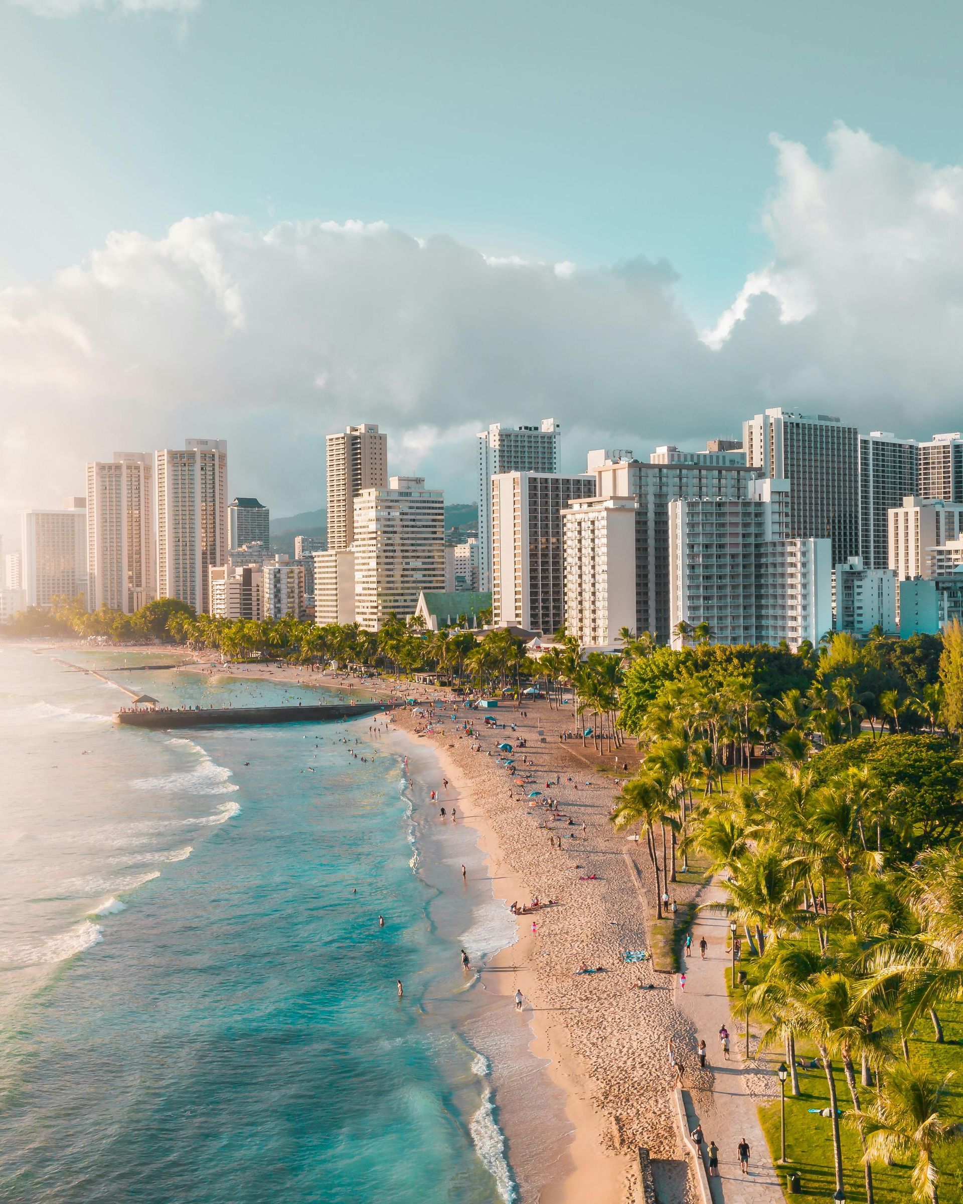 Hawaii cityscape with beach, ocean, palm trees, and high-rise buildings under a cloudy sky.