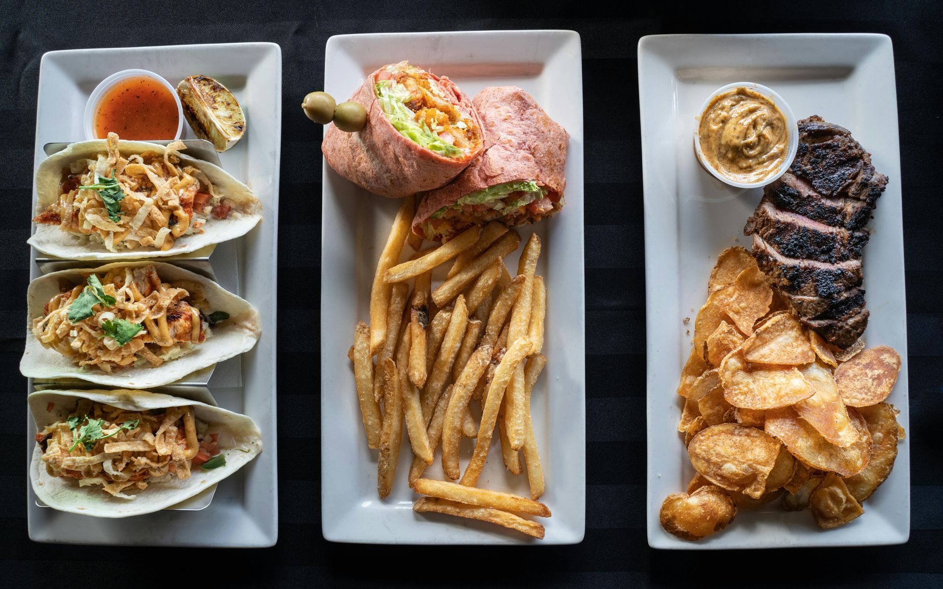 Three rectangular plates with various food: tacos, wrap with fries, steak with chips and dipping sauce.