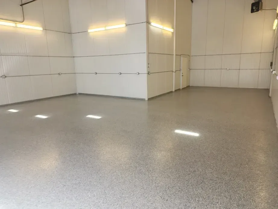 Empty room with gray speckled floor and white walls, lit by long overhead lights.