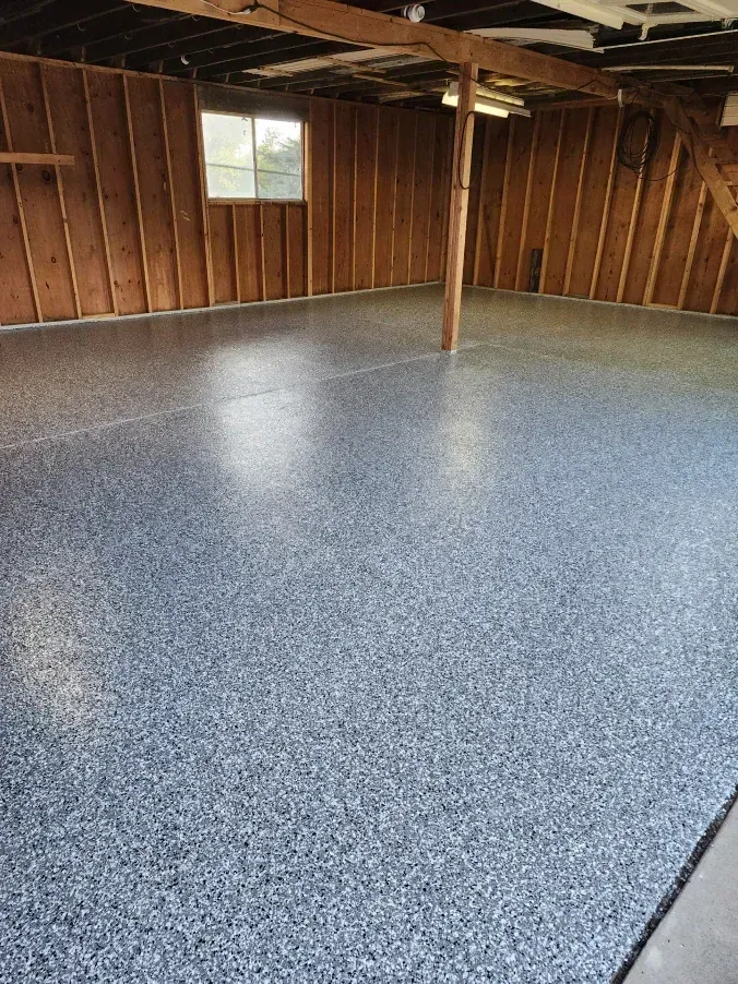 A garage with a newly coated speckled floor, wooden walls, a small window, and support beams.