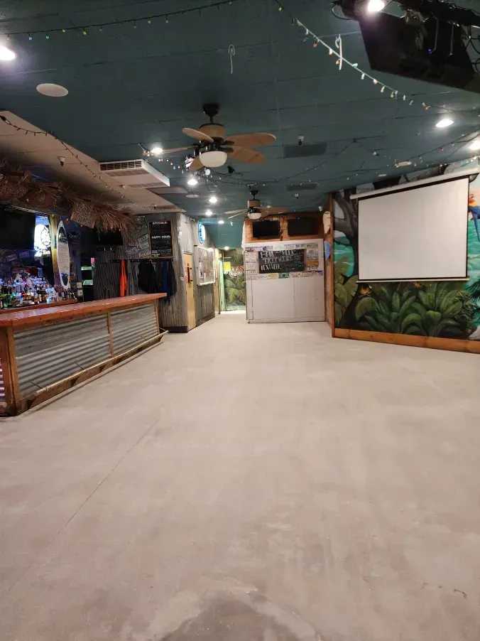 Empty interior of a bar with a corrugated metal bar front, jungle-themed murals, and a projector screen.