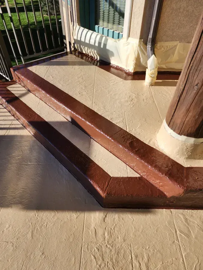 Brown-painted steps and pillar on a beige textured concrete surface, near a window and railing.
