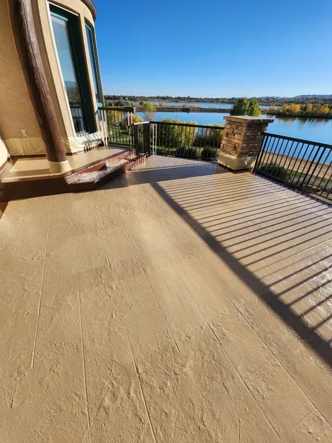 Patio overlooking water, sunny day. Tan-colored floor, dark railing and view of distant shore.