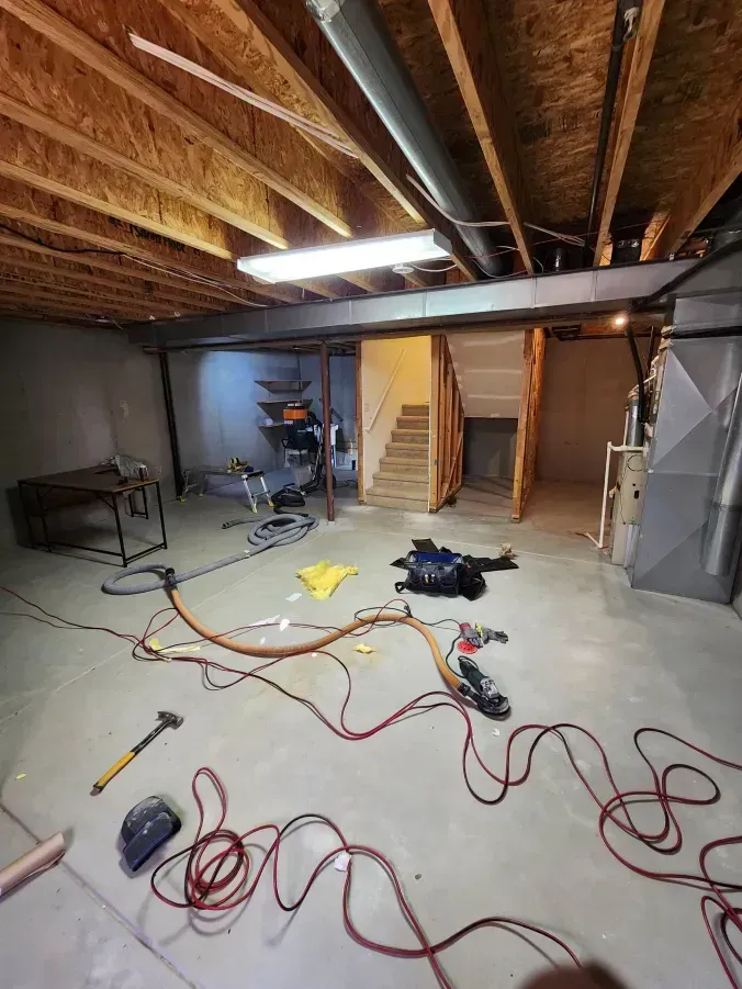 Basement under construction, concrete floor, exposed ceiling joists, electrical wires strewn about. Staircase leads up.