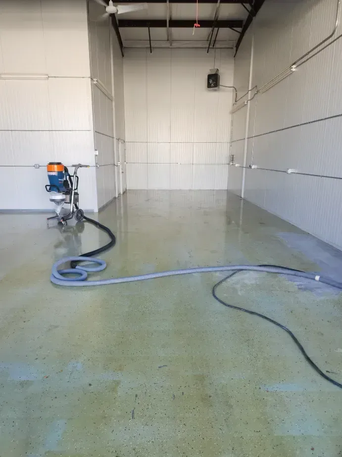 Interior of a large, empty room with a floor grinder and hoses. Walls are white and lined. Floor is wet.