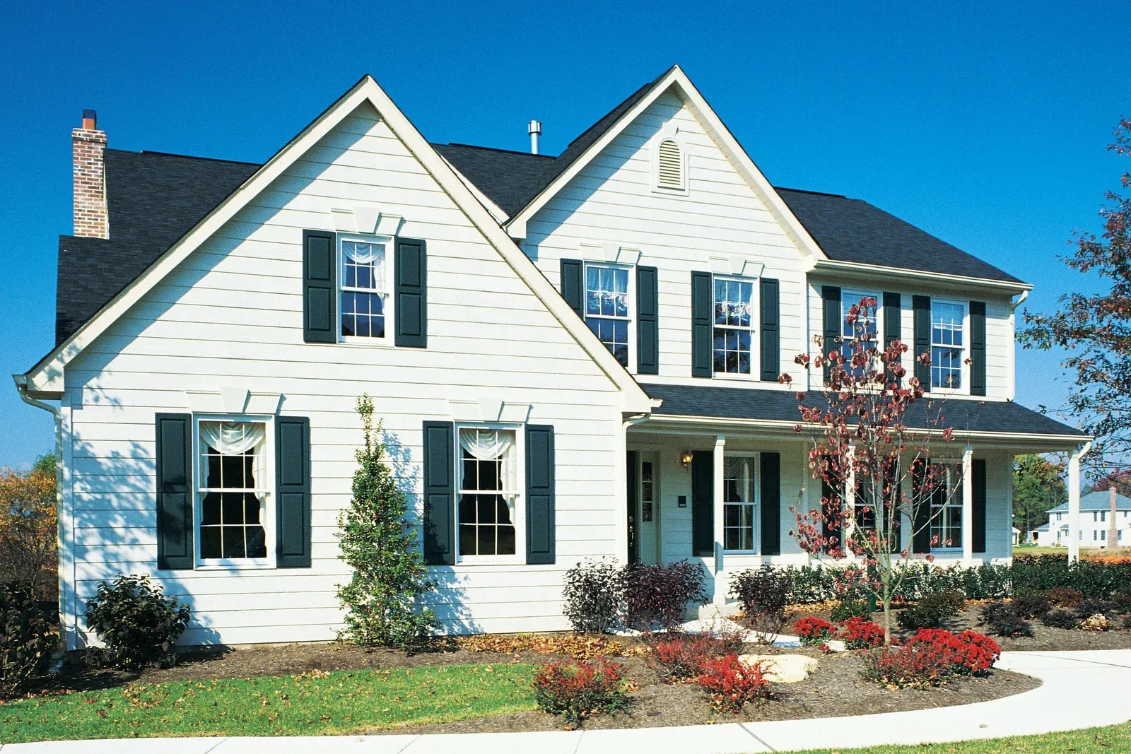 White two-story house with black shutters, chimney, and dark roof against a clear blue sky.