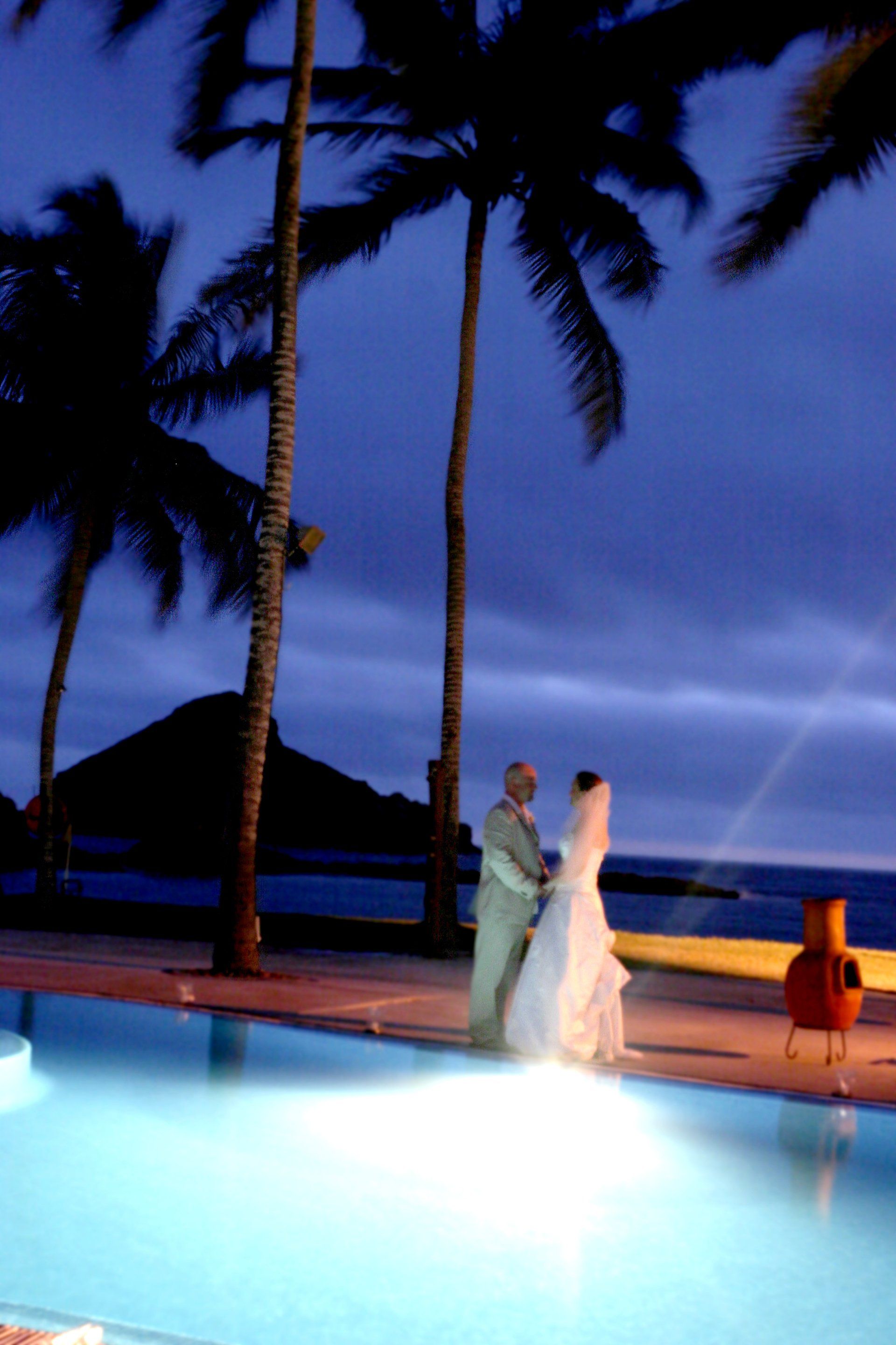 A bride and groom standing next to a swimming pool at night
